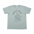 A light sage green graphic t-shirt laid flat on a brown cardboard surface. The shirt features a dark gray graphic of a person riding a bicycle with the text 'ATWATER' and 'EST.