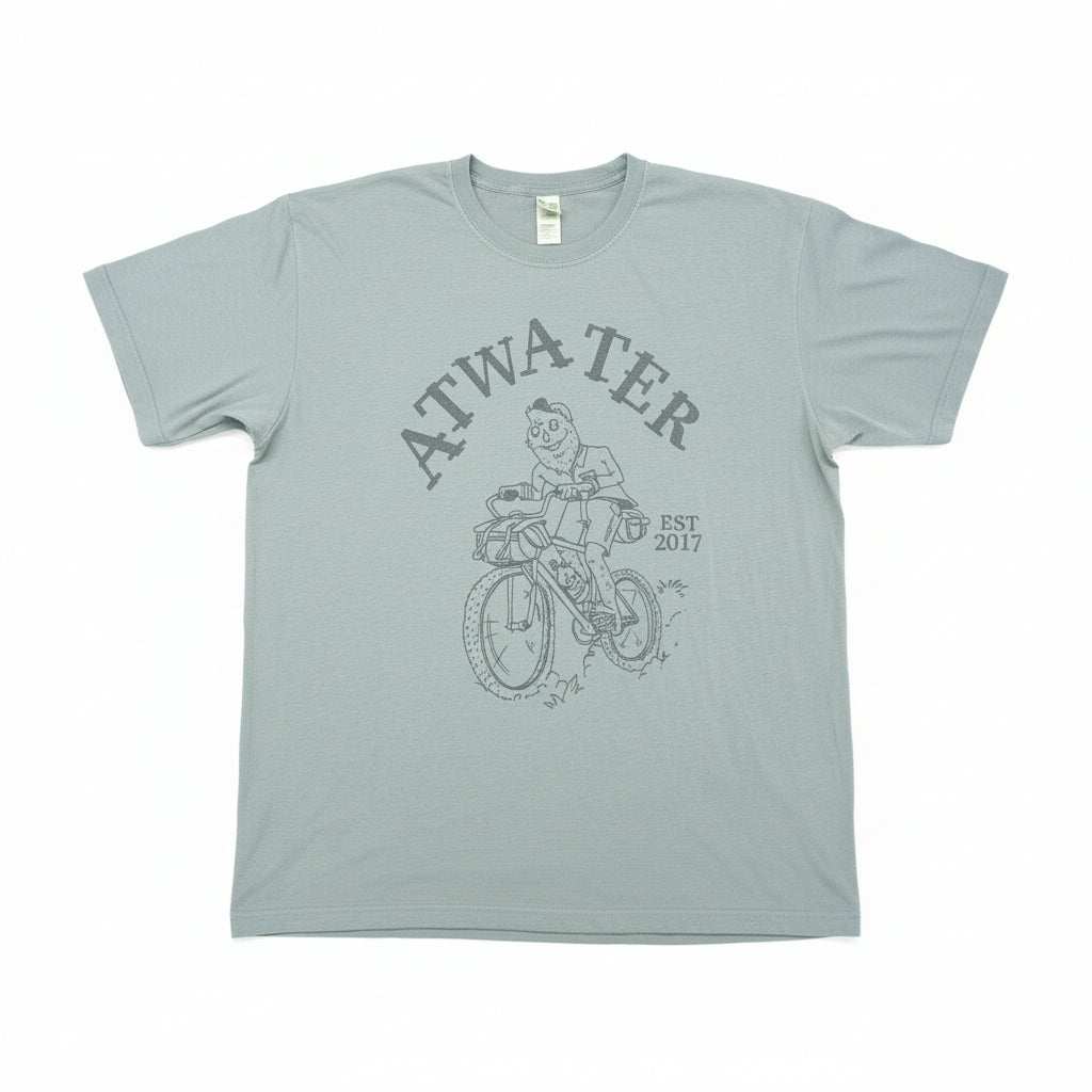 A light sage green graphic t-shirt laid flat on a brown cardboard surface. The shirt features a dark gray graphic of a person riding a bicycle with the text 'ATWATER' and 'EST.